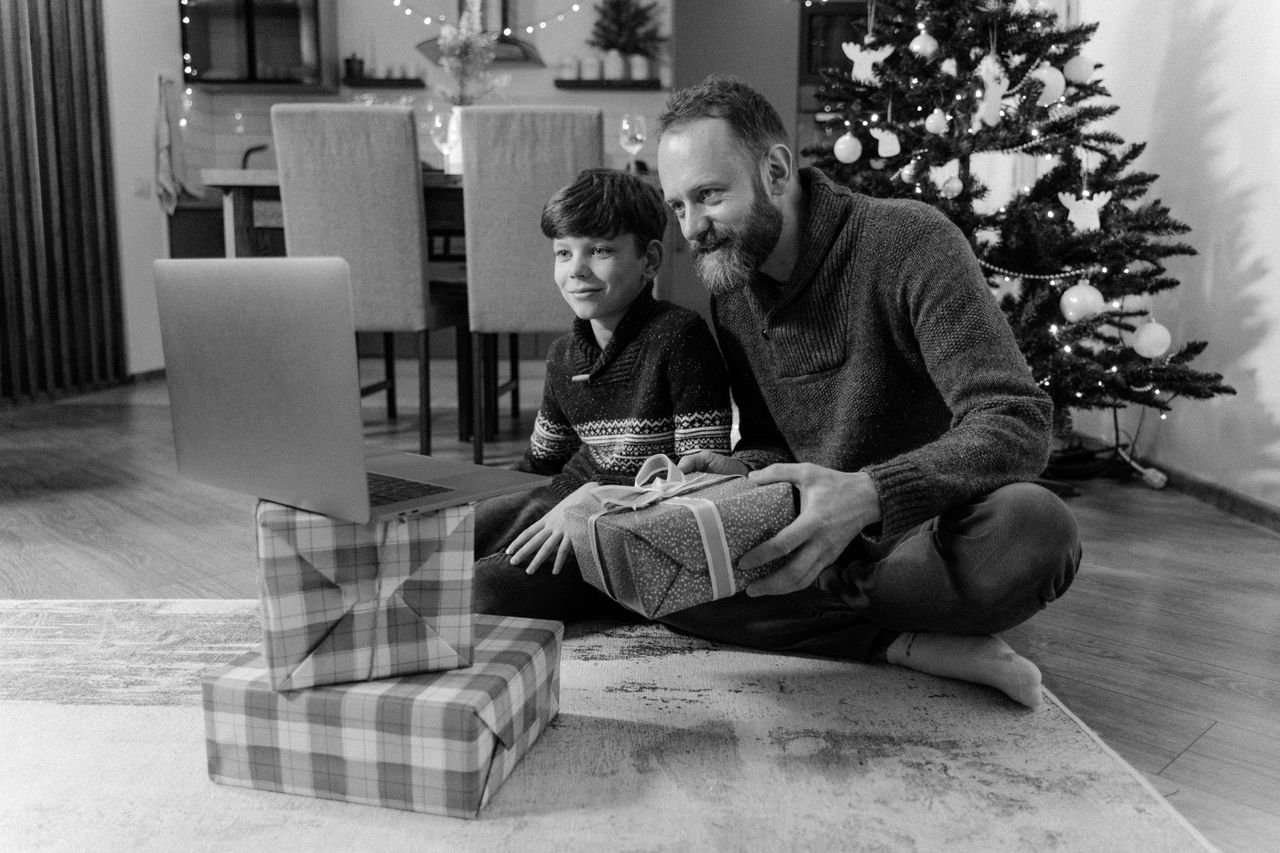 services-04 Father and son sitting by Christmas tree video calling relatives and holding gifts.