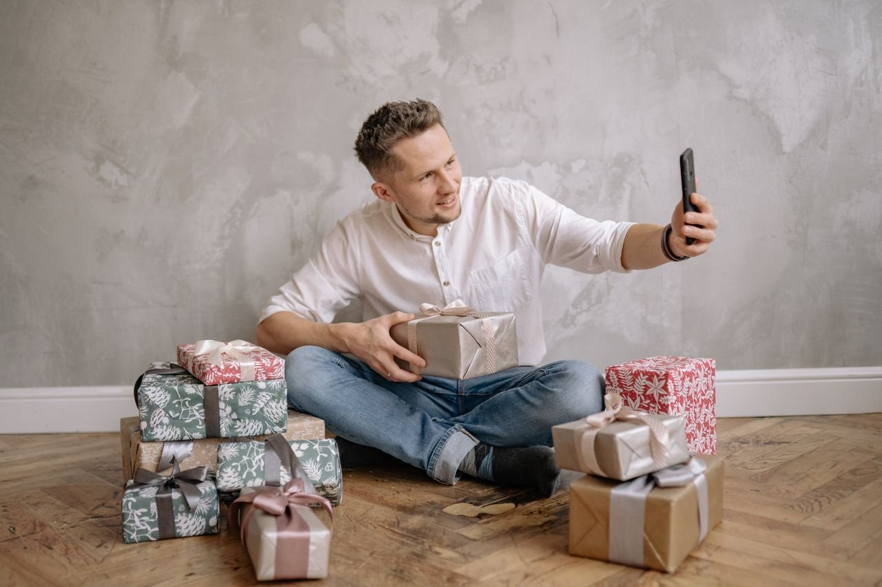 heros-img A man sitting on the floor surrounded by wrapped presents, taking a selfie with his smartphone.